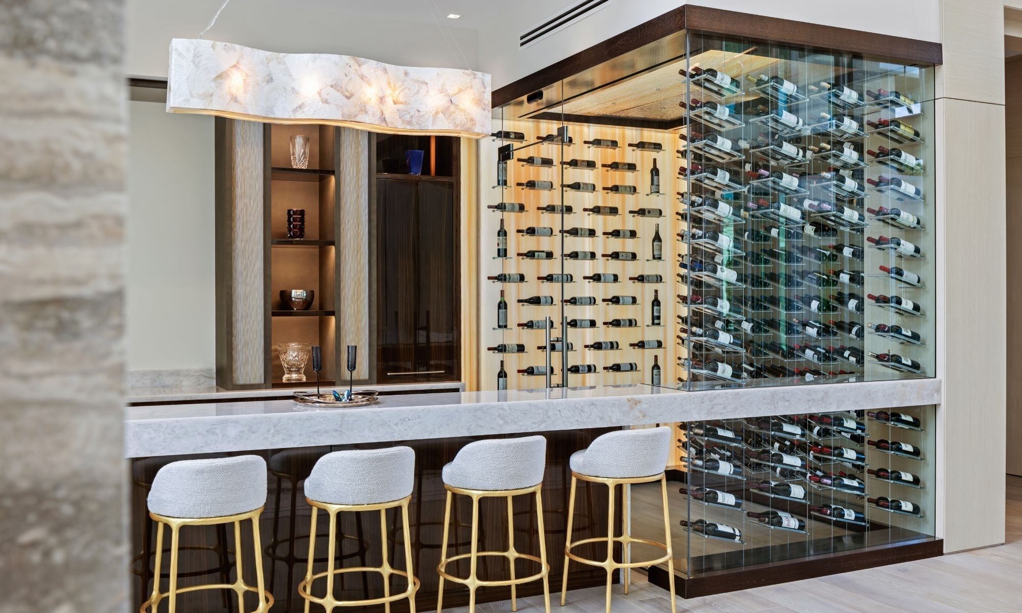 Modern glass wine cellar with backlit racks beside a marble bar and stylish stools