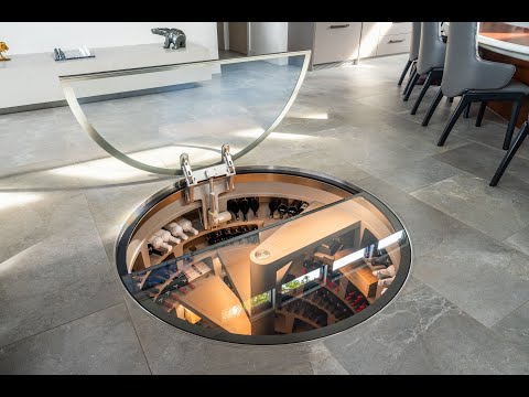 A high-angle view of a Spiral Cellar subterranean wine storage system, featuring a signature concrete spiral staircase surrounded by integrated wine racking. The image showcases the architectural integration into the home's floor with a luxury glass hatch